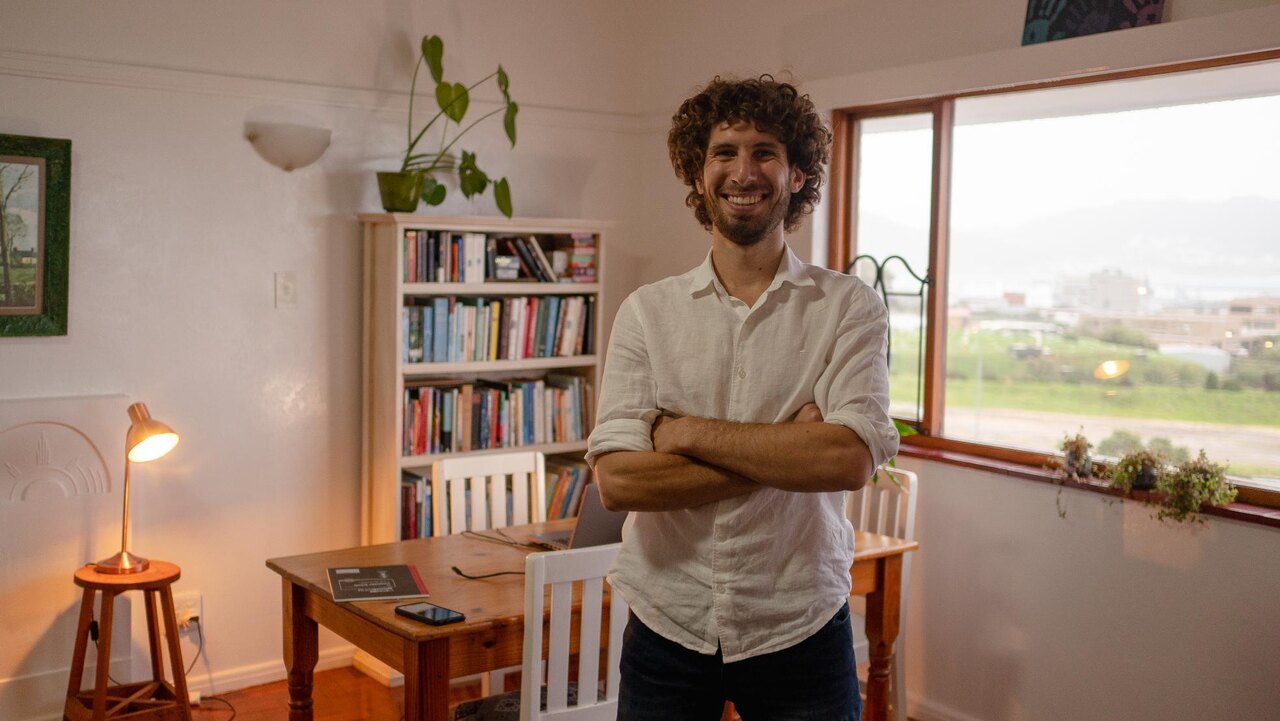 John Naiker, founder of Think Camp, standing with arms folded in a warm, book-lined room overlooking Cape Town.